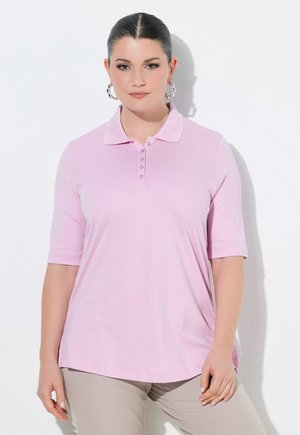 Kobieta o ciemnych włosach w jasnoróżowej koszulce polo z krótkim rękawem i beżowych spodniach, stojąca na białym tle, zwrócona twarzą do przodu.