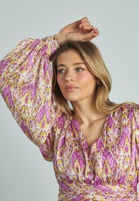 Blusa multicolorida con un vibrante patrón abstracto en púrpura y dorado, fabricada en tejido ligero, con mangas largas abullonadas y escote en V.