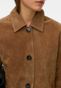 Pruun seemisnahast jakk lõdva lõikega, millel on krae ja mustad nupud. Tekstuur on pehme ja veidi läikiv.