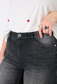 Jeans in denim nero con una finitura sbiadita, caratterizzati da un design classico a cinque tasche e una chiusura con bottone. Abbinati a una camicia bianca con ricamo di cuori rossi.