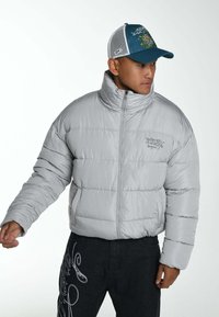 Grijze pufferjack met een hoge kraag en horizontale stiksels, gedragen over zwarte broek met witte grafische accenten. De pet is teal met een mesh achterkant.