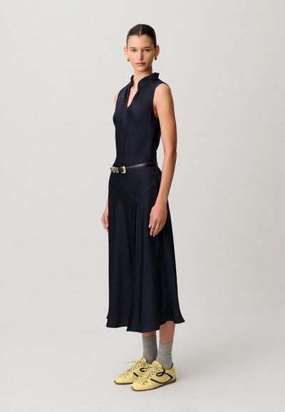 Mujer con un vestido midi negro sin mangas con cinturón, calcetines grises, zapatillas amarillas y pendientes de aro dorados, posando frente a un fondo liso.