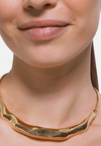 Goldfarbener Halsreifen mit wellenförmigem, unregelmäßigem Design. Glatte Oberfläche mit glänzendem Finish, auf einem nackten Hals ruhend. Einzigartige, skulpturale Form.
