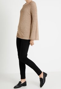 Beżowy sweter z dzianiny o teksturowanym przodzie i dzwonowymi rękawami, zestawiony z czarnymi spodniami slim fit i czarnymi loafersami.
