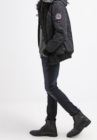 Giacca nera con cappuccio e bordo in pelliccia, felpa grigia sotto, jeans skinny scuri e stivali neri stringati. Toppa dettagliata sulla manica.