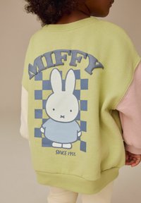 Grafički dukserica s zelenom pozadinom, oversized dizajn, rukavi u ružičastoj i krem boji te plavi Miffy lik s natpisom.
