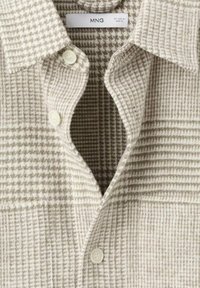 Camisa beige de cuadros con botones, confeccionada en una tela texturizada, que cuenta con cuello, tres botones y un bolsillo, hecha de un material suave y ligero.