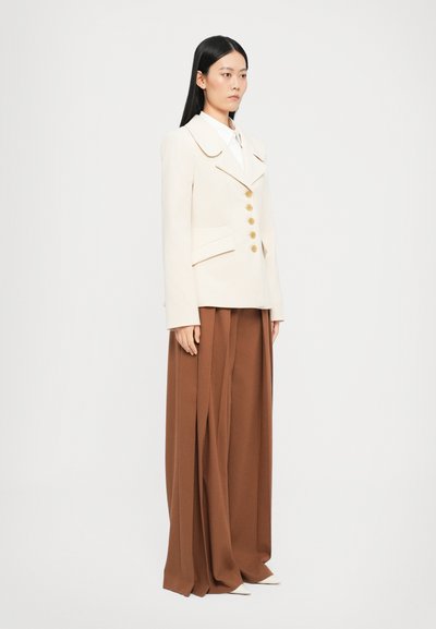 Blazer crema con un amplio cuello y botones dorados, combinado con pantalones marrones con pliegues. Presenta un diseño ajustado y una textura suave.