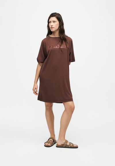 Femme debout, les bras derrière le dos, portant une robe marron ample au niveau des genoux avec manches courtes et logo, et des sandales marron sur fond blanc.