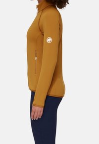 Giacca aderente con zip e maniche lunghe in giallo senape, realizzata in tessuto morbido; presenta una tasca laterale e un logo circolare sulla manica.