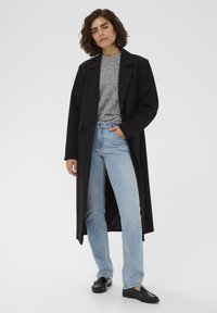 Femme debout portant un long manteau noir, un pull gris à motifs, un jean bleu clair et des mocassins noirs, sur un fond blanc uni.