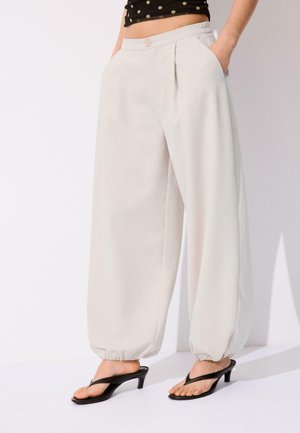 Kremowe spodnie o wysokim stanie i szerokich nogawkach z elastycznymi ściągaczami, noszone z czarnymi sandałami na obcasie i czarnym krótki topem w białe kropki.