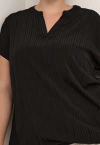 Haut noir à manches courtes avec un décolleté en V, présentant une texture côtelée et un léger brillant. Le tissu semble léger et lisse.