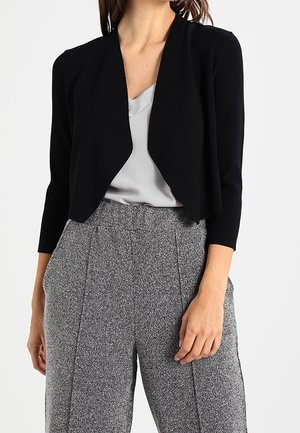 Femme portant un cardigan court noir, un chemisier gris et un pantalon gris texturé taille haute avec coutures avant et poches.