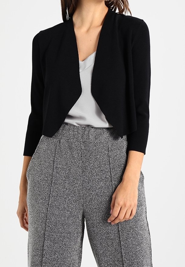 Femme portant un cardigan court noir, un chemisier gris et un pantalon gris texturé taille haute avec coutures avant et poches.
