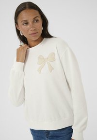 Sweatshirt en tissu crème doux avec un nœud beige brodé sur la poitrine, avec des poignets et une bande de taille côtelés, coupe décontractée.
