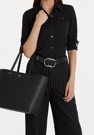 Femme portant une chemise noire à boutons et un pantalon noir à fines rayures avec ceinture à boucle argentée, tenant un grand sac fourre-tout en cuir noir avec un logo doré.