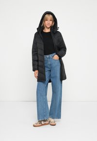 Manteau noir rembourré avec capuche, présentant une fermeture éclair et des poches latérales, associé à un jean bleu à jambes larges et des baskets à motifs.