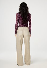 Langärmlige lila, florale, durchsichtige Bluse mit figurbetontem Schnitt, kombiniert mit hellbeigen Schlaghosen mit hinteren Taschen und leichter Naht.
