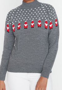 Maglione grigio lavorato a maglia con una fila di piccoli pupazzi di Babbo Natale rossi e bianchi sul petto e sulle spalle, indossato con pantaloni bianchi.