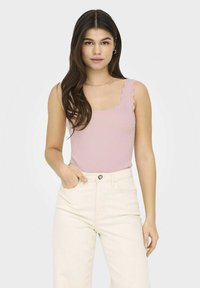 Top sin mangas de color rosa claro con bordes en forma de concha, combinado con jeans de tiro alto en color blanco roto. El modelo tiene el cabello largo y las manos en los bolsillos.