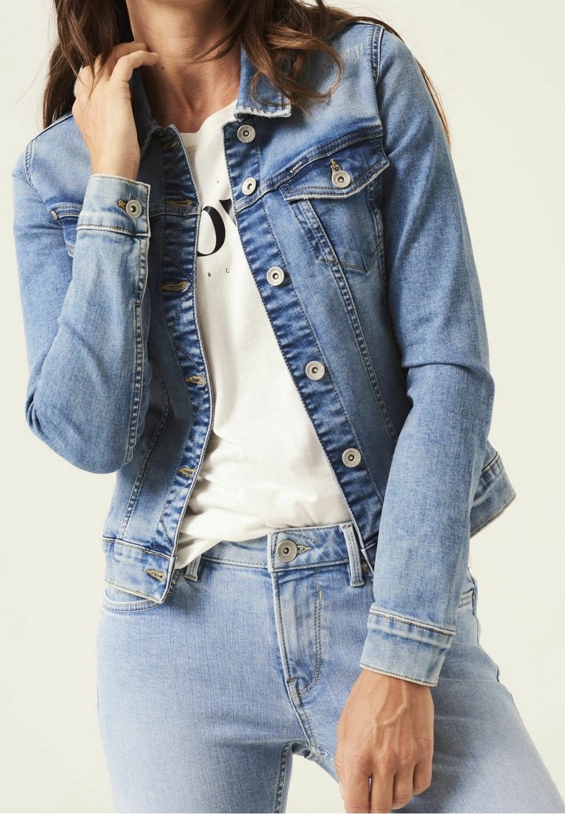 Vrouw in een lichtblauwe denimjack over een wit shirt met zwarte tekst, gecombineerd met bijpassende lichtblauwe denim jeans.