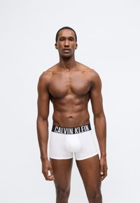 Λευκά ανδρικά εσώρουχα boxer με μαύρη ελαστική μέση που φέρει το λογότυπο "CALVIN KLEIN". Ο τόνος του δέρματος είναι μεσαίος, με ομαλή υφή και εφαρμοστό σχεδιασμό.