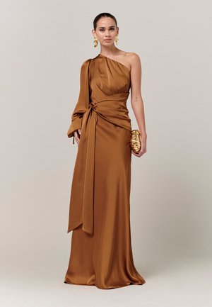 Woman wearing a one-shoulder, long brown satin gown with a tied waist sash, holding a gold clutch and wearing gold leaf-shaped earrings.