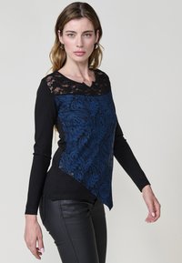 Haut à manches longues avec une partie supérieure en dentelle noire et une petite moitié inférieure à motif paisley bleu, avec un ourlet asymétrique et des accents de boutons.