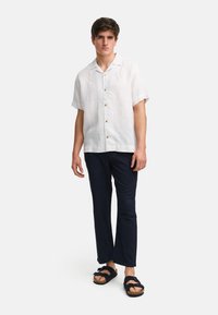 Camisa de lino blanca, ligera y de manga corta, con botones de madera, combinada con pantalones oscuros de color marino y sandalias negras con tiras gruesas.
