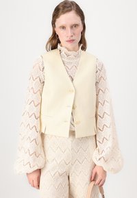 Gilet beige chiaro con tre bottoni, indossato sopra una blusa in pizzo con colletto alto. I pantaloni texture presentano un motivo in pizzo dettagliato.