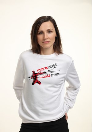Kvinde med skulderlangt brunt hår iført en hvid Marvel Studios sweatshirt med Deadpool, der holder sværd.
