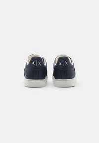 Sneaker in pelle blu navy con suola bianca, caratterizzati da un design minimalista e un logo embossato sul tallone. Texture liscia e forma arrotondata.