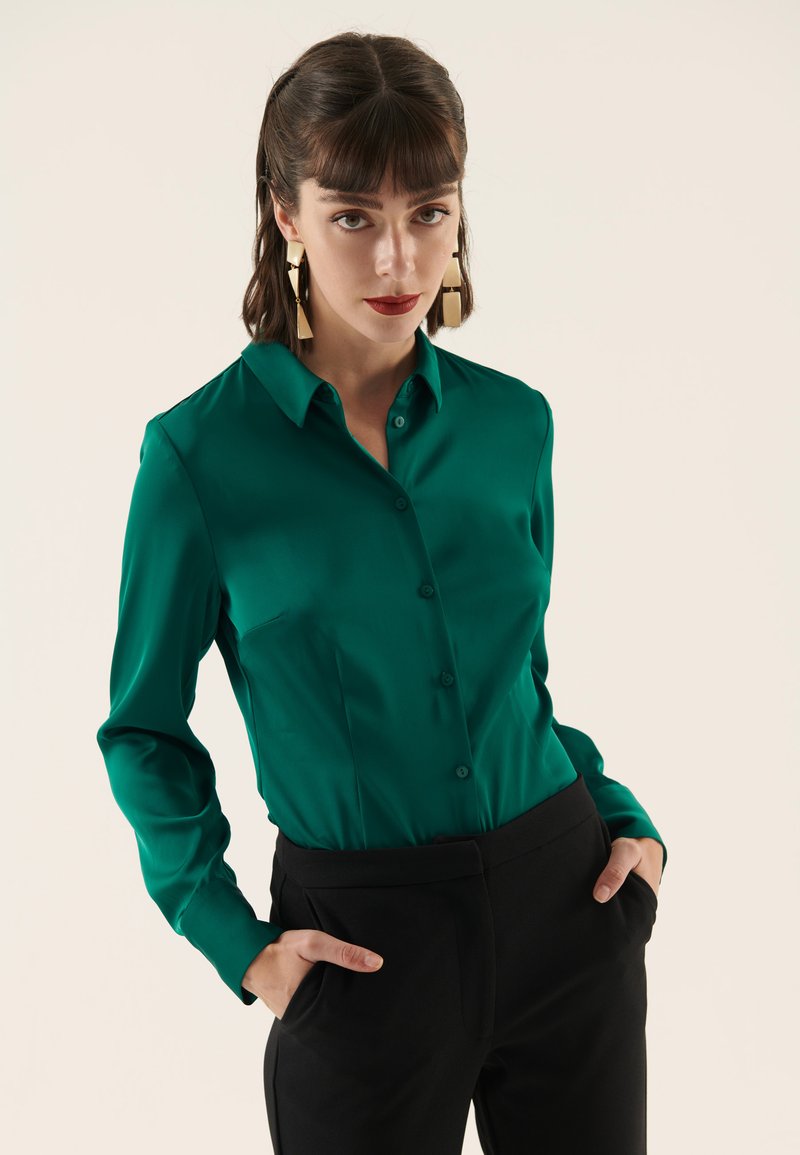 Blusa in raso verde con colletto, chiusura con bottoni e maniche lunghe, abbinata a pantaloni neri a vita alta.