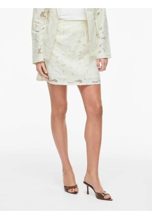 Kvinne iført hvit blomsterblonde skjørt, matchende jakke og brune åpen-tå hælmulesko, stående foran en ensfarget bakgrunn.