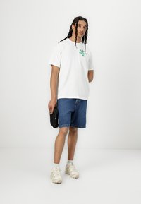 White cotton t-shirt with green graphic print, paired with blue denim shorts and beige sneakers. Holds a small black bag.