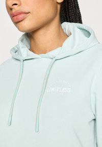 Φούτερ με κουκούλα σε ανοιχτό μέντα πράσινο με λευκό λογότυπο κειμένου. Διαθέτει κορδόνι και απαλό υλικό. Η τσέπη τύπου καγκουρό δεν είναι ορατή. Απλός σχεδιασμός.