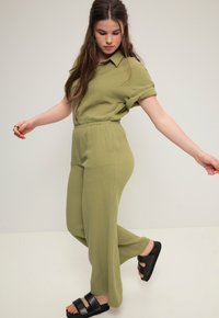 Olivgrünes Zweiteiler-Outfit mit einem kragenlosen Oberteil, umgeschlagenen Ärmeln und relaxed-fit Hose. Strukturierter Stoff; getragen mit schwarzen Sandalen.