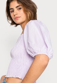 Top lilás com padrão xadrez, mangas bufantes, corpete pregueado e decote elástico, apresentando uma bainha com babados e um tecido texturizado.