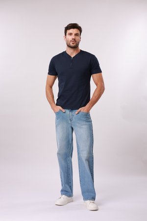 Mand stående med hænderne i lommerne iført en mørkeblå kortærmet skjorte, lyseblå jeans og hvide sneakers mod en ensfarvet baggrund.