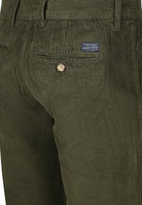 Close-up of olive green corduroy pants with a buttoned back pocket and a blue fabric brand label.