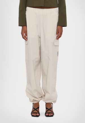 Pantalon cargo beige avec poignets élastiques porté par une personne avec une veste courte vert olive et des sandales noires à brides sur fond blanc.
