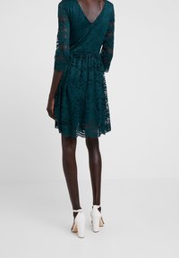 Türaqua Spitzenkleid mit Dreiviertelärmeln, V-Ausschnitt hinten, ausgestelltem Rock und gewelltem Saum. Kombiniert mit weißen High Heels.