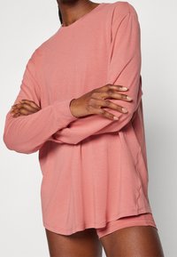 Langärmliges, rosa Oberteil aus weichem Stoff mit Rundhalsausschnitt und lockerem Schnitt, kombiniert mit passenden Shorts. Sichtbare Nähte und ein Seitenschlitz-Detail.