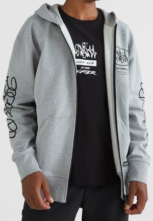 Uomo che indossa una felpa con cappuccio grigio chiaro con cerniera, con testo grafico nero sulle maniche e sul petto, sopra una t-shirt nera con stampa di testo bianco.