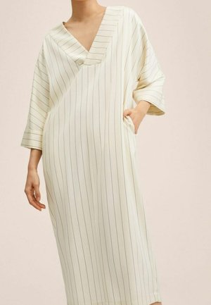 Femme portant une robe ample couleur crème avec de fines rayures verticales et un profond décolleté en V, posant avec une main dans la poche.