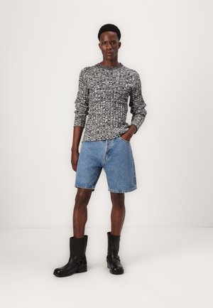Pull en tricot à motifs gris et noir, shorts en denim bleu clair, bottines noires à boucle. La modèle se tient face à l'avant.