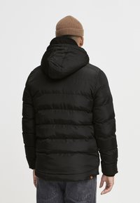 Mann in schwarzer gepolsterter Jacke mit Kapuze und braunem Strickmütze, der mit dem Rücken zu einem schlichten hellen Hintergrund steht.