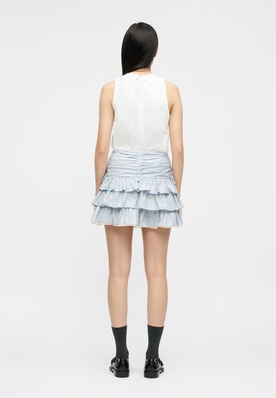 Γυναίκα με μακριά σκούρα μαλλιά, που φοράει λευκό αμάνικο τοπ, στρωτή φούστα με ανοιχτό μπλε βολάν, σκούρες κάλτσες και μαύρα μοκασίνια, στέκεται με την πλάτη προς τα πίσω.