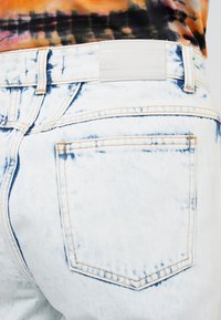 Světle modré džíny z denimu s opotřebovanou texturou, oranžovým prošíváním a bílou koženou nášivkou s nápisem "CLOSED" na pasu.